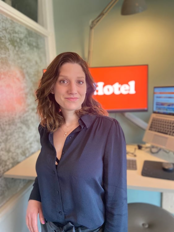 Smiling professional woman in an office setting with a large monitor displaying the word 'Hotel' and a laptop on a modern desk.