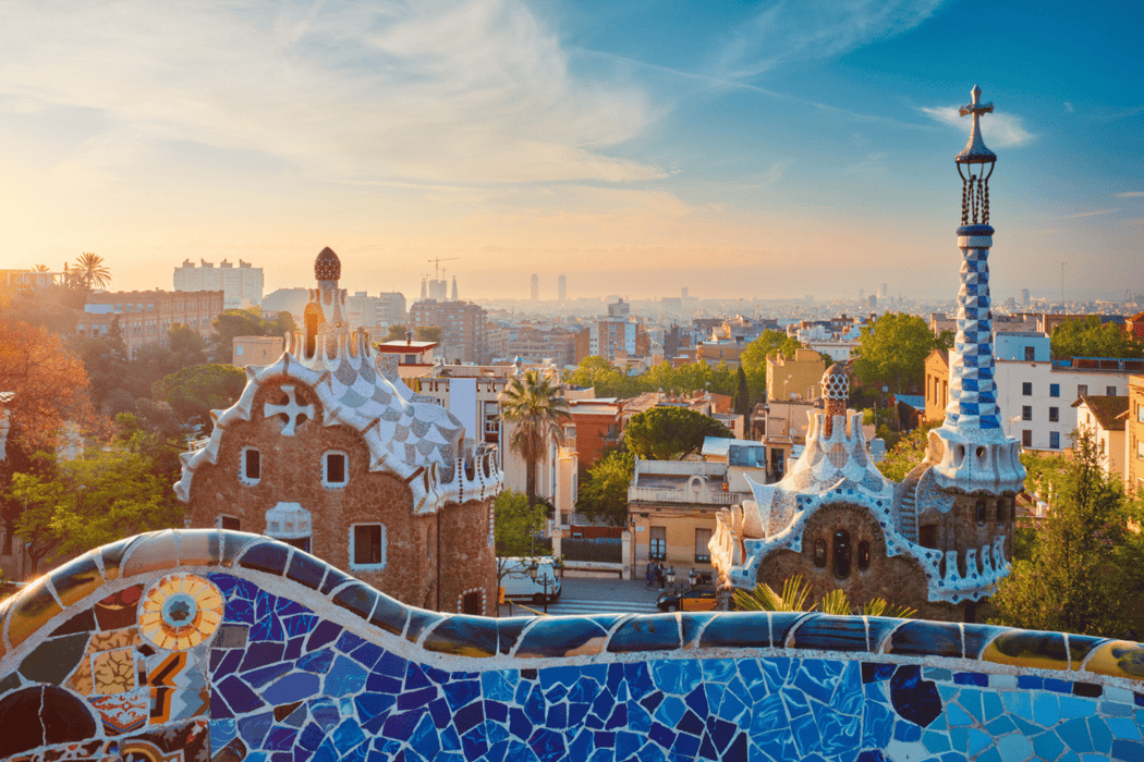 Vibrant sunrise panorama of Barcelona's Park Güell, showcasing the iconic mosaic-tiled serpentine bench, whimsical Gaudí houses, and the city skyline under a golden sky.