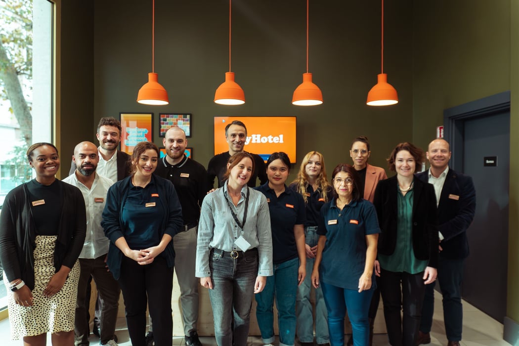A diverse team of easyHotel employees, including front-desk staff and management, smiling in a modern easyHotel lobby. The easyHotel logo is visible on a screen, complemented by signature orange light fixtures.