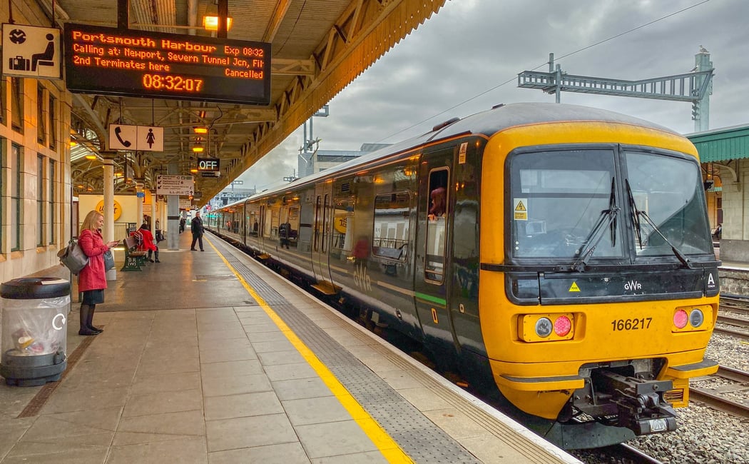 A yellow and black Great Western Railway (GWR) Class 166 train awaits departure at Bristol Temple Meads station. Passengers stand on the platform, and a digital display board shows a cancelled service to Portsmouth Harbour via Newport and Severn Tunnel Ju