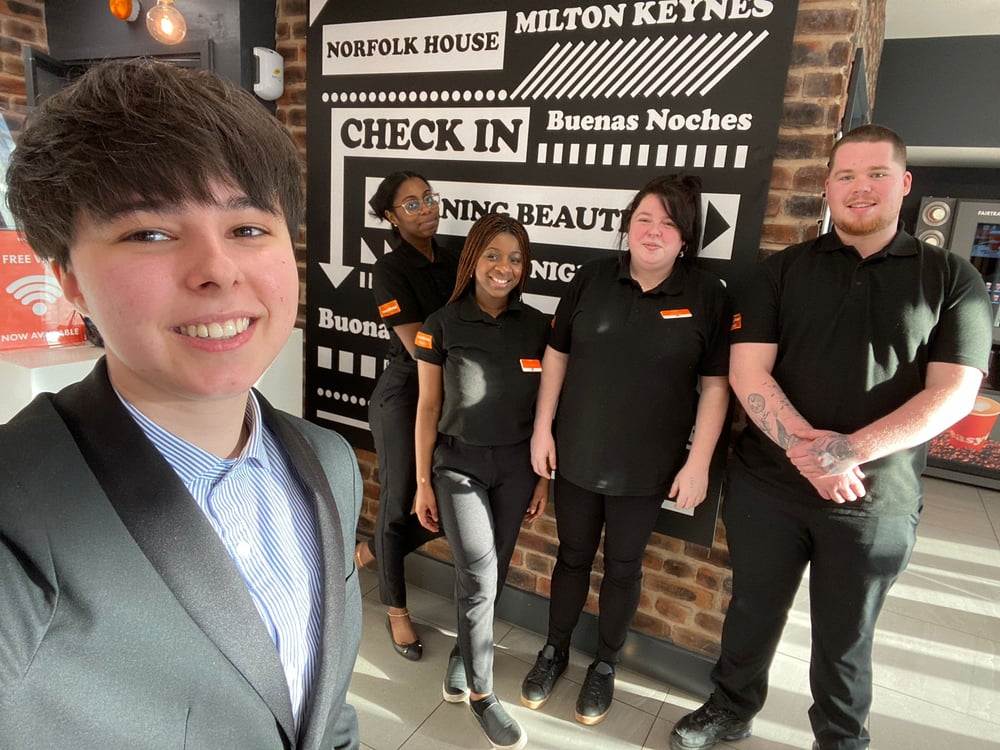 A smiling team of five easyHotel staff, wearing black uniforms with orange accents, stand in the modern reception area of easyHotel Milton Keynes, with a large graphic sign displaying 'MILTON KEYNES', 'NORFOLK HOUSE', and 'CHECK IN' visible behind them.