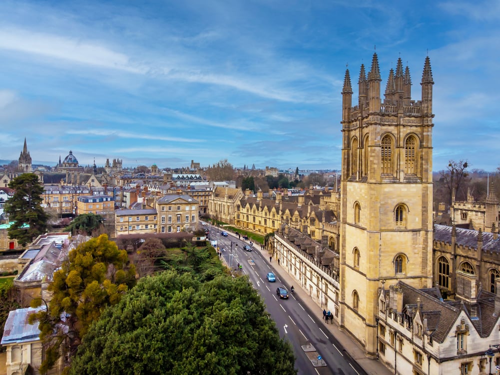 Aerial view of Oxford city skyline featuring iconic university buildings including Magdalen College Tower, historic colleges, a street with cars and pedestrians, and green trees under a partly cloudy sky.