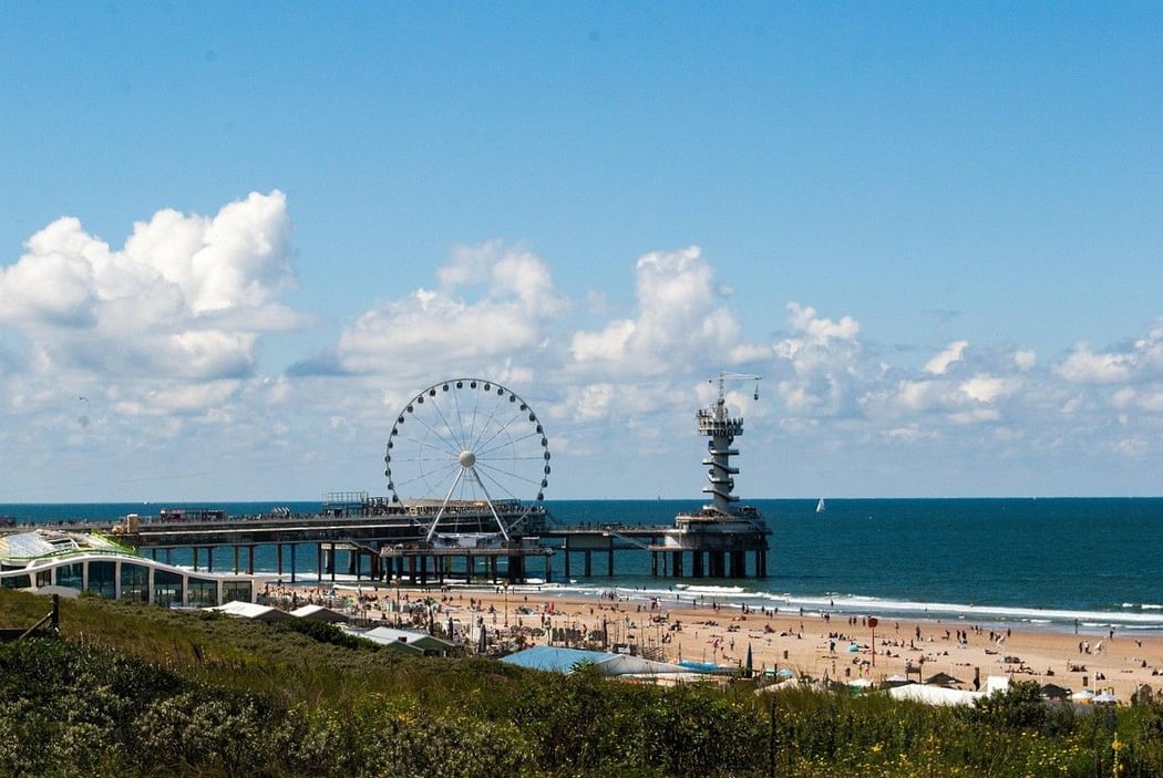 Panoramic view of Scheveningen beach and pier in The Hague, Netherlands, on a sunny day. The iconic pier features a large Ferris wheel and a bungee jumping tower, with people relaxing on the sandy shore.