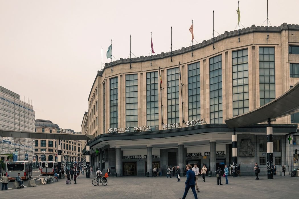 Brussels Central Station exterior, featuring its curved, stone Art Deco facade with tall windows. The bustling plaza below is filled with pedestrians and a bus, depicting a lively urban scene in Brussels, Belgium.