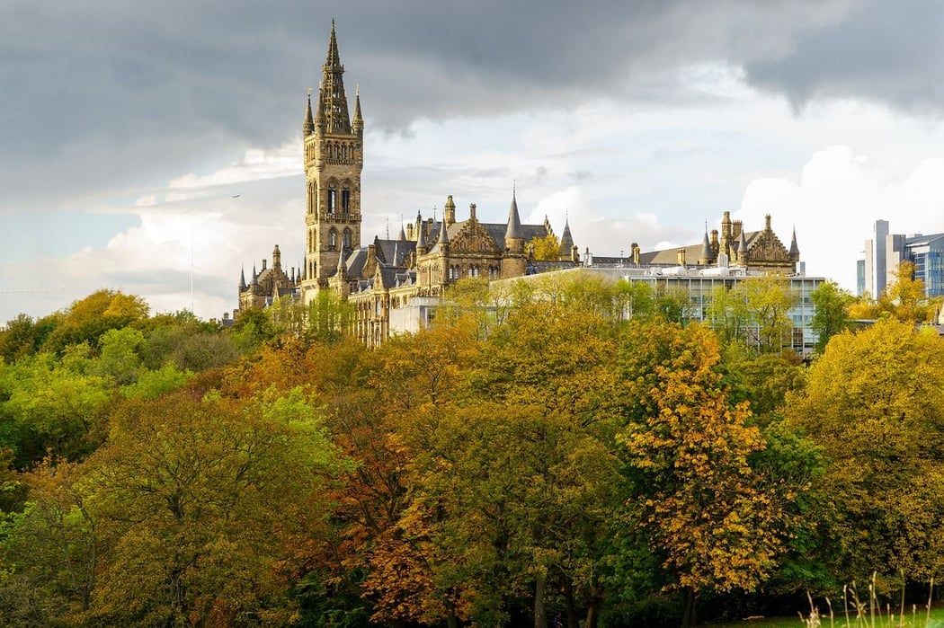 The iconic University of Glasgow main building, featuring its tall Gothic tower, rises above a vibrant canopy of yellow and orange autumn trees in Glasgow, Scotland.