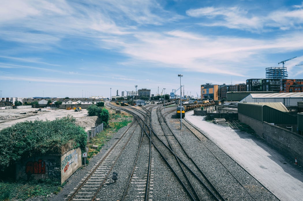 Overhead view of railway tracks converging in a train yard in Dublin, Ireland, with a yellow locomotive, industrial port cranes, and modern city buildings under a bright blue sky.