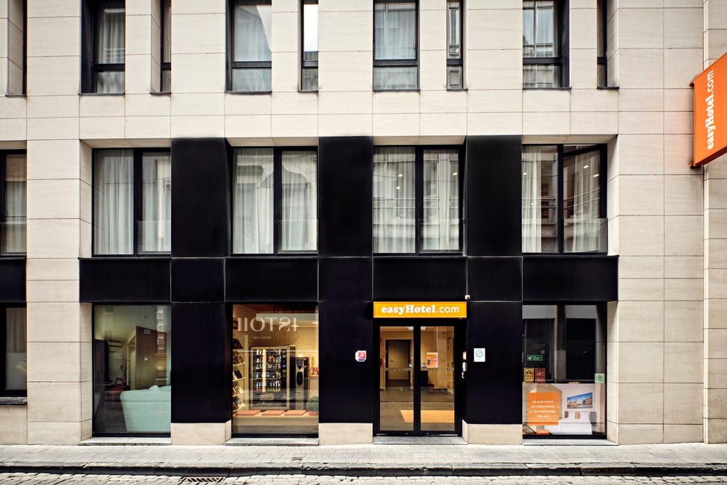Front exterior of the easyHotel Brussels City Centre, a modern building with contrasting black and light-colored panels, featuring the prominent easyHotel branding, street-level windows, and an entrance on a cobblestone street.
