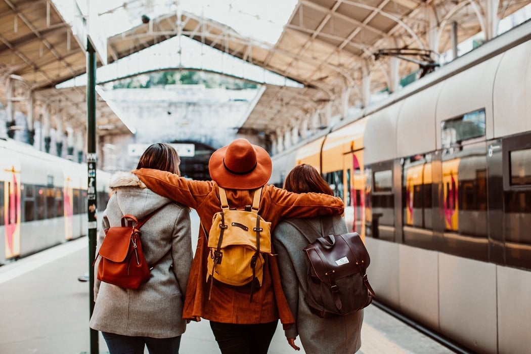Three friends with backpacks stand arm-in-arm on a European train station platform with modern trains, ready for travel.