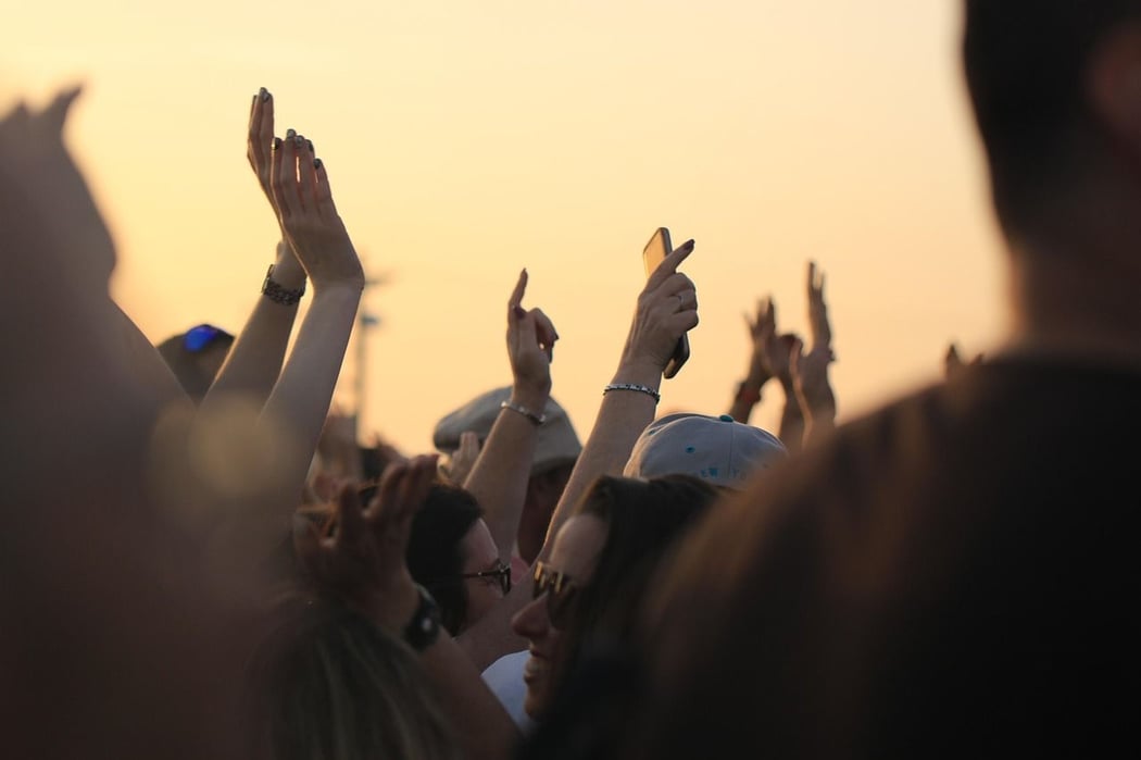 Outdoor concert crowd with hands raised and phones held high, silhouetted against a warm, golden sunset sky.