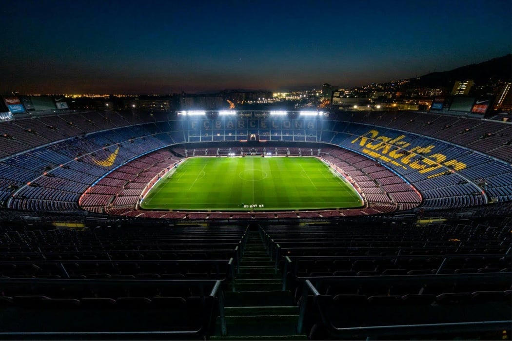 High-angle view of the empty Camp Nou football stadium in Barcelona at night, featuring the brightly lit green pitch and illuminated 'FC Barcelona' text on the stands.