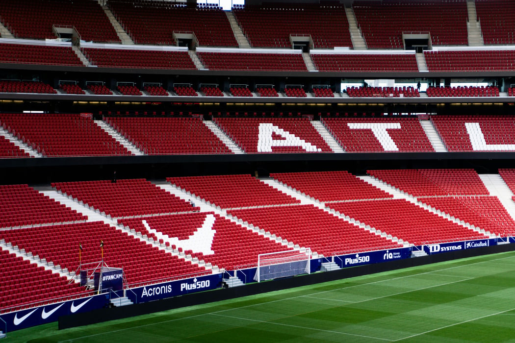 An empty Cívitas Metropolitano stadium in Madrid, Spain, featuring rows of red seats, with white seats forming "ATLÉTICO" in the upper tier and a large club crest in the lower tier, overlooking a green football pitch.