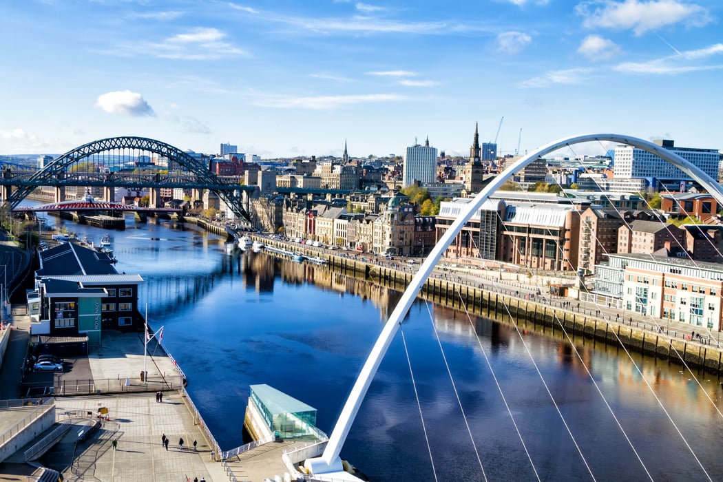 Panoramic view of Newcastle upon Tyne cityscape overlooking the River Tyne, featuring the iconic Tyne Bridge and Millennium Bridge, with historic buildings lining the bustling quayside under a clear blue sky.