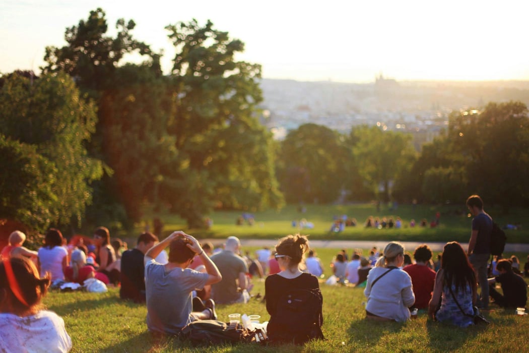 People relaxing on a grassy hillside in a Prague park, overlooking the city skyline and St. Vitus Cathedral at sunset.