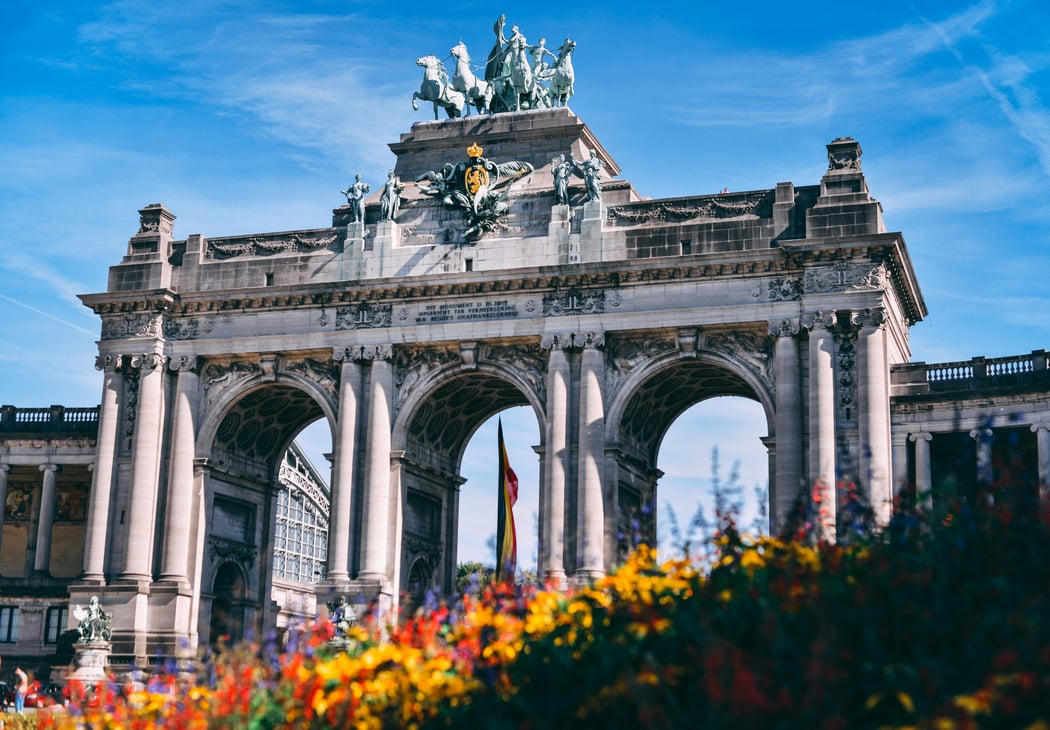 Cinquantenaire Arch in Brussels, Belgium, a historic triumphal arch with a quadriga statue and intricate sculptures, seen through a foreground of vibrant colorful flowers under a clear blue sky.