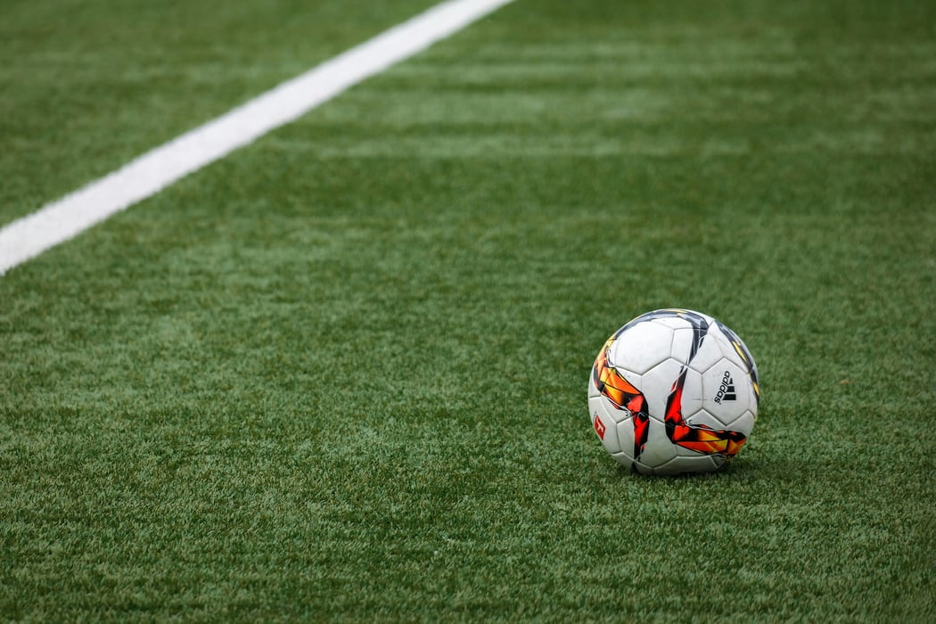 A white, orange, and red Adidas soccer ball rests on a green artificial turf football pitch with a white boundary line.