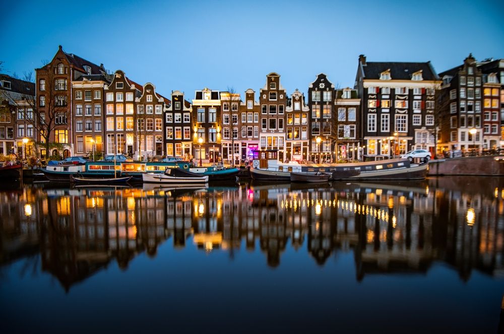A scenic twilight view of historic Amsterdam canal houses and houseboats illuminated along a tranquil canal, with their reflections shimmering on the water.