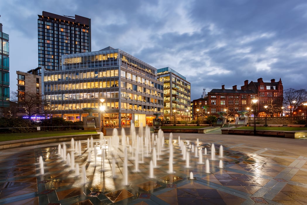 Dusk cityscape of Piccadilly Gardens in Manchester, UK, featuring an illuminated water fountain in the foreground. Modern glass office buildings with lit windows stand prominently behind, alongside a distinctive historic red-brick building under a cloudy 