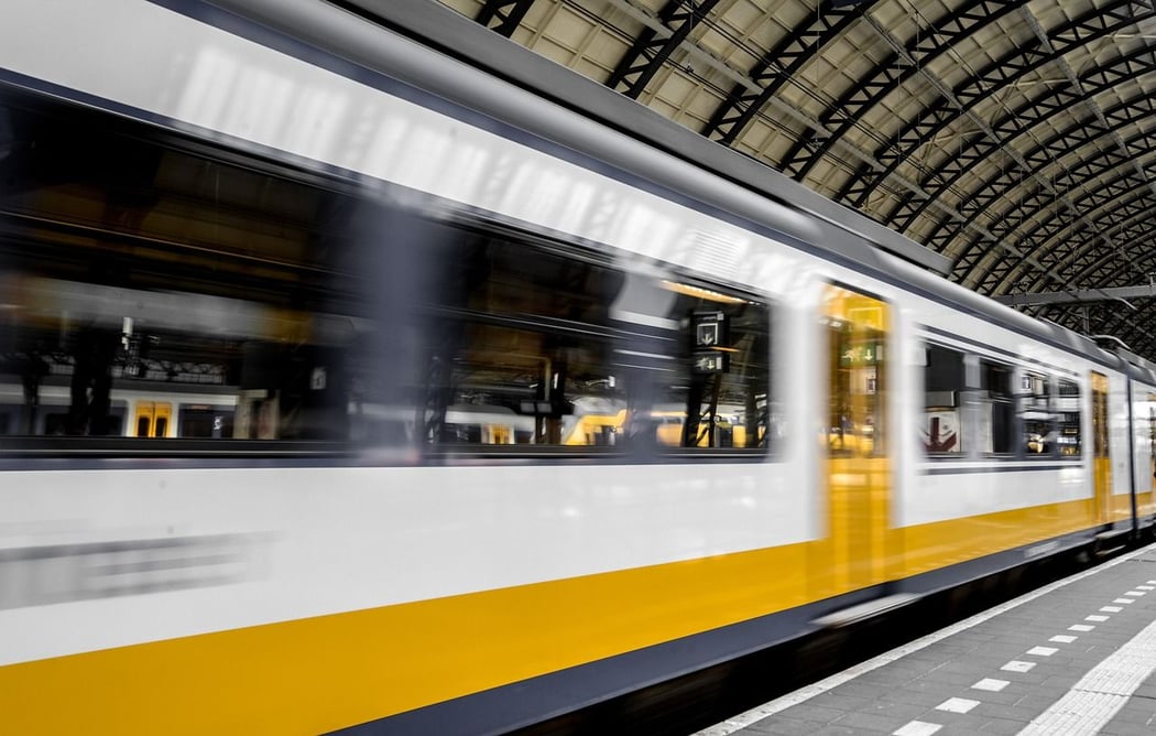 High-speed passenger train, white with a yellow stripe, captured with motion blur as it moves through Amsterdam Centraal Station, featuring its iconic arched roof.