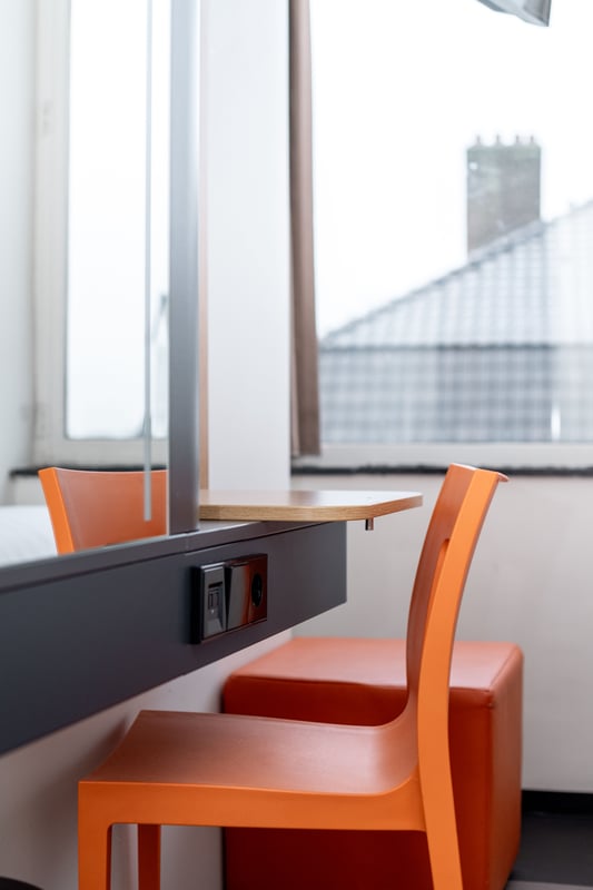 A minimalist desk area in an easyHotel Maastricht room, featuring a bright orange chair, a built-in wooden shelf, and convenient power outlets, with a view of rooftops through the window.
