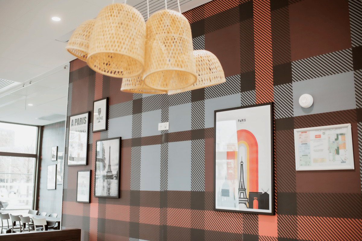 Modern common area at easyHotel Paris Nord Aubervilliers, featuring stylish wicker pendant lights above a wall with distinctive brown, grey, and orange plaid wallpaper, decorated with various framed Paris-themed artworks.