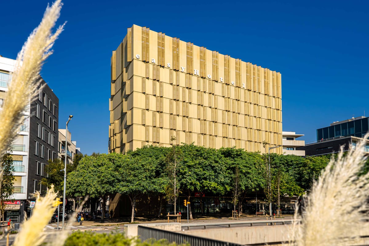 Exterior view of the modern easyHotel building in Barcelona, featuring its light brown facade with numerous windows and vertical "easyHotel" signage, seen from a green park with tall ornamental grasses and trees under a clear blue sky.