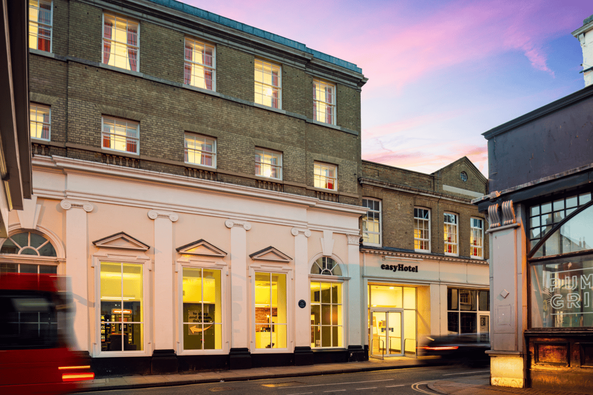 Exterior view of easyHotel Ipswich at dusk, featuring the illuminated street-level entrance and windows of the brick building with classical details under a vibrant pink and purple sky.