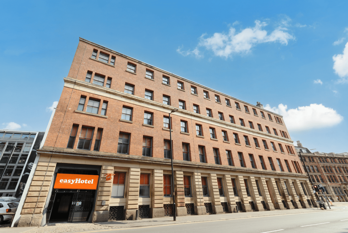 Exterior view of the easyHotel Manchester building, a multi-story brick hotel with a prominent orange easyHotel sign at street level under a clear blue sky.