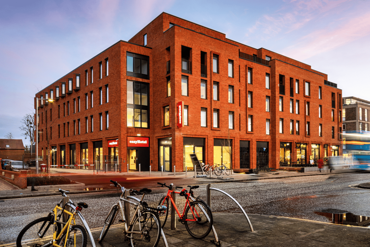 easyHotel Oxford's modern red-brick building exterior at dusk, featuring illuminated windows and prominent branding. The street scene includes bicycles parked on a wet pavement and a passing bus with motion blur, suggesting a lively city environment.