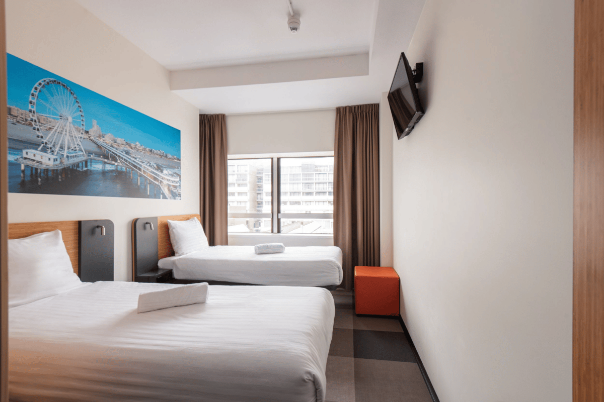 Modern easyHotel Scheveningen Beach room with two single beds, brown curtains, wall-mounted TV, and a large mural depicting the Scheveningen Pier with its Ferris wheel.