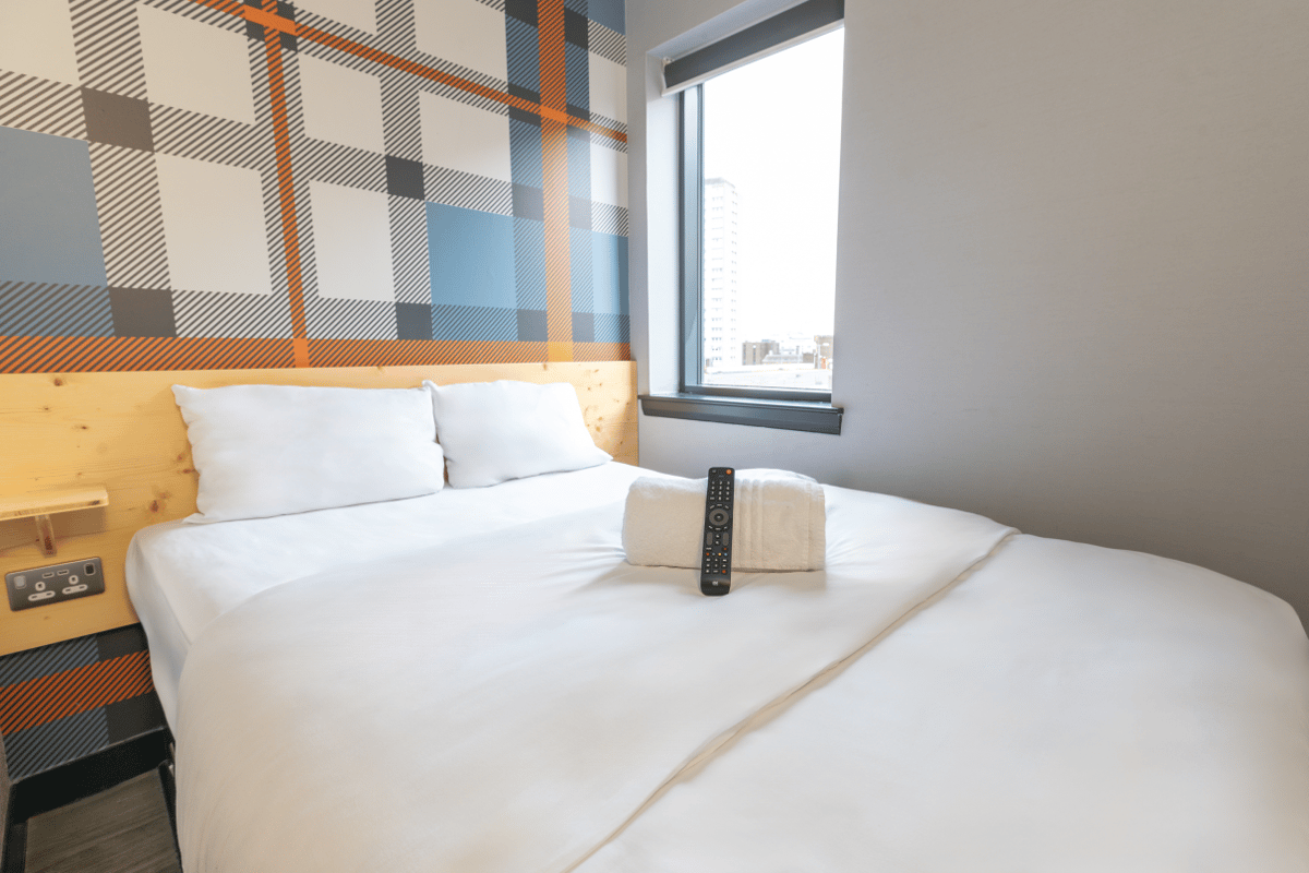 ALT: easyHotel Glasgow room with a comfortable bed, white linens, vibrant tartan accent wall, and city window view.