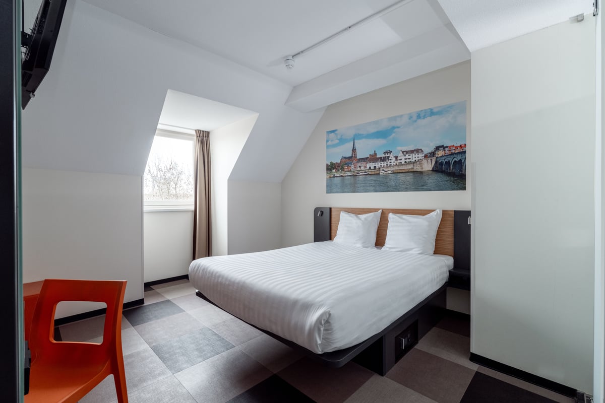 A modern easyHotel room in Maastricht featuring a large bed with white linens, checkered flooring, and a vibrant mural of Maastricht's cityscape, including the Maas River and bridge, above the headboard. The room has a bright window and minimalist design.