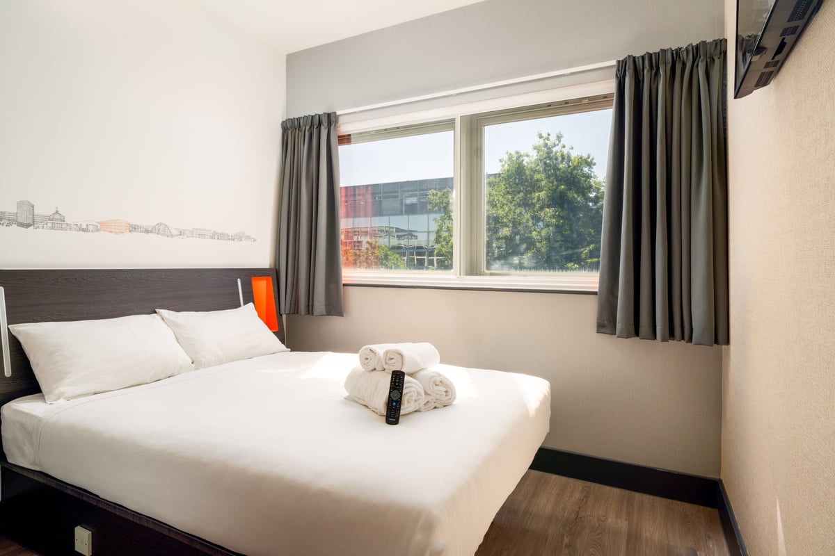 A modern and bright easyHotel room in Milton Keynes, featuring a double bed with crisp white linens, a stack of clean white towels, and a TV remote. A large window with grey curtains offers a view of trees and a building, while a city skyline mural decora