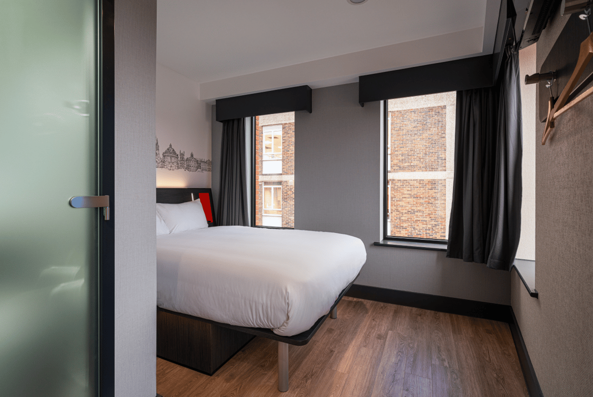 easyHotel Oxford room with a comfortable double bed, white bedding, and a black and white Oxford cityscape mural above the bed. The modern room features two large windows, dark grey walls, and light wooden flooring.