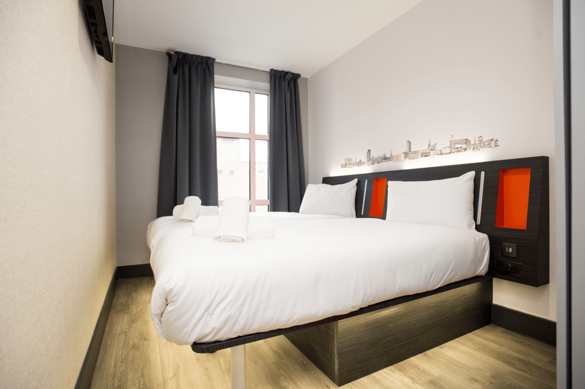 Modern easyHotel Sheffield room featuring a double bed with white linen, pillows, and rolled towels. The room has a dark grey headboard with orange accents, a Sheffield cityscape mural above the bed, light wood-effect flooring, and dark grey curtains by a