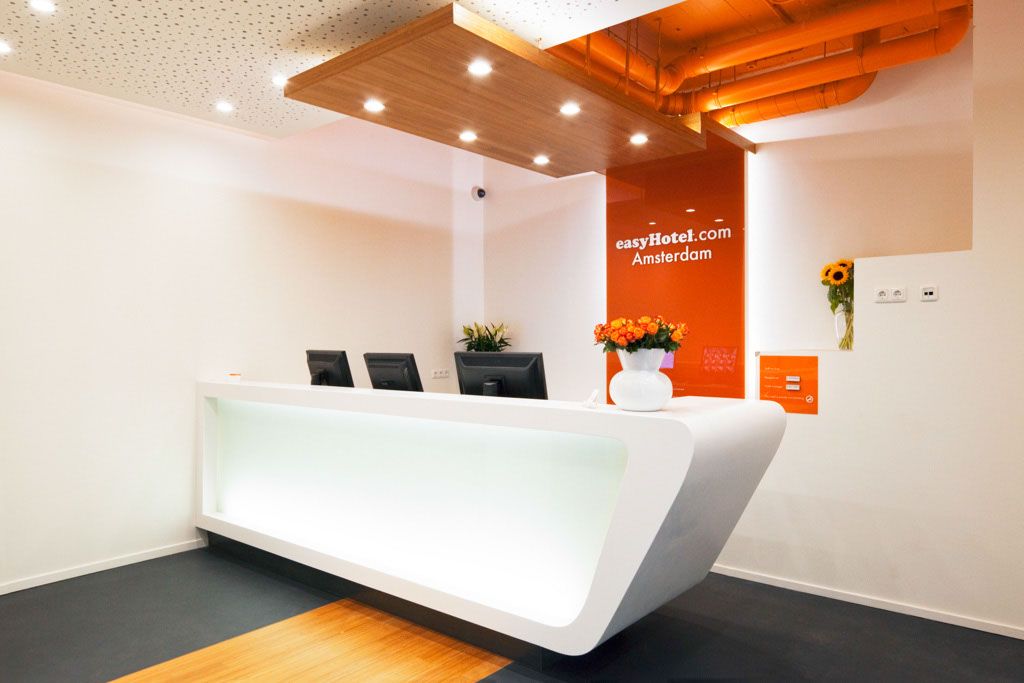 Bright and modern reception desk at easyHotel Amsterdam City Centre South. The lobby features a sleek white counter, orange branded wall panel, and vibrant floral decor, highlighting the hotel's minimalist design.