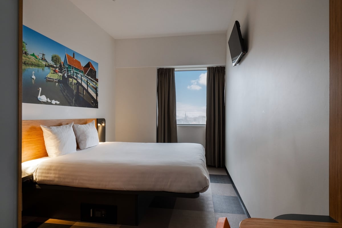 Modern easyHotel Amsterdam Zaandam room with a neatly made bed, a large wall art print of a traditional Dutch canal scene with windmills and swans, a window offering an urban view, and a wall-mounted TV.