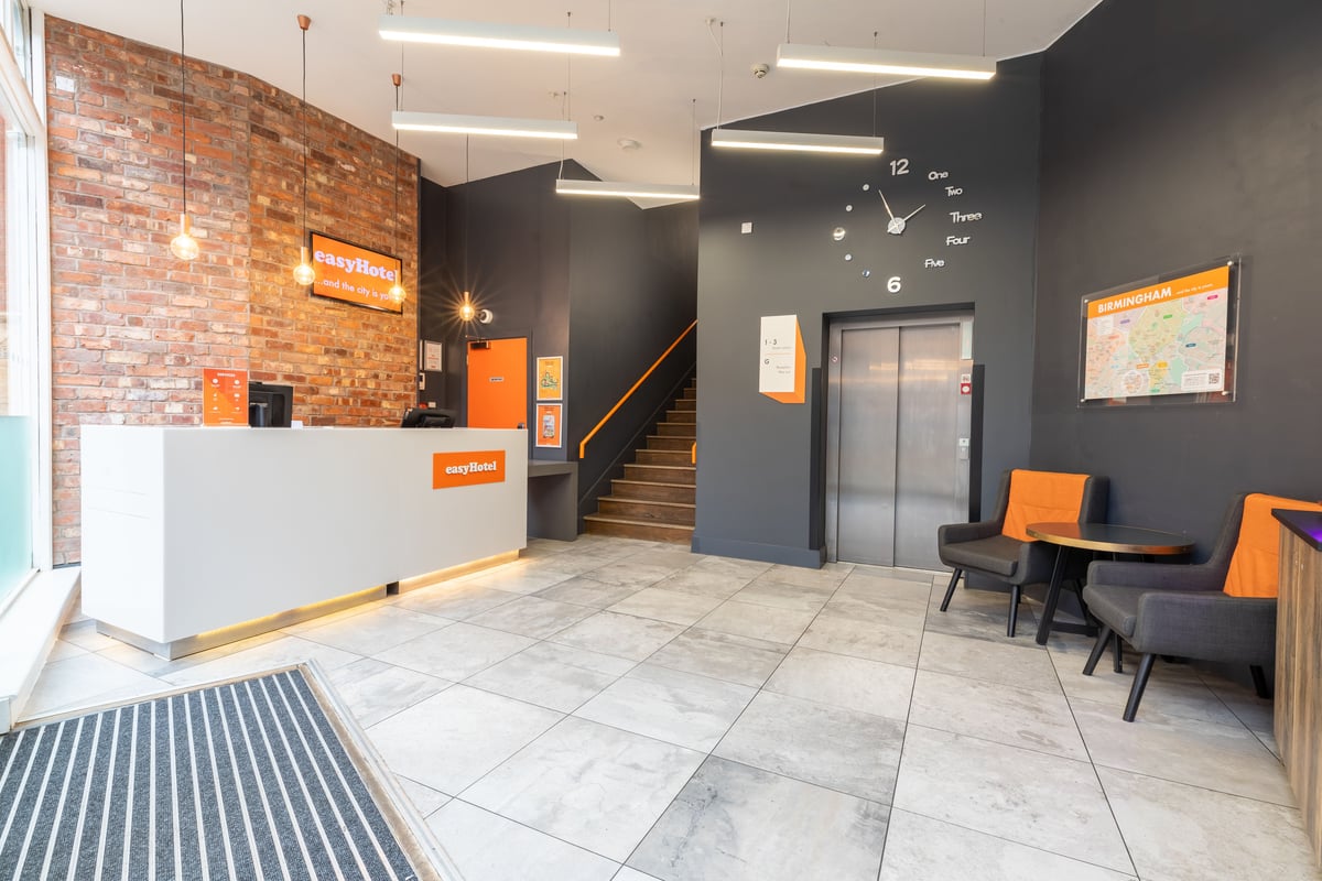 A bright, modern easyHotel Birmingham lobby featuring a white reception desk with an easyHotel logo, exposed brick wall, and dark grey walls with orange accents. The space includes a comfortable seating area, a large wall clock, an elevator, and stairs le