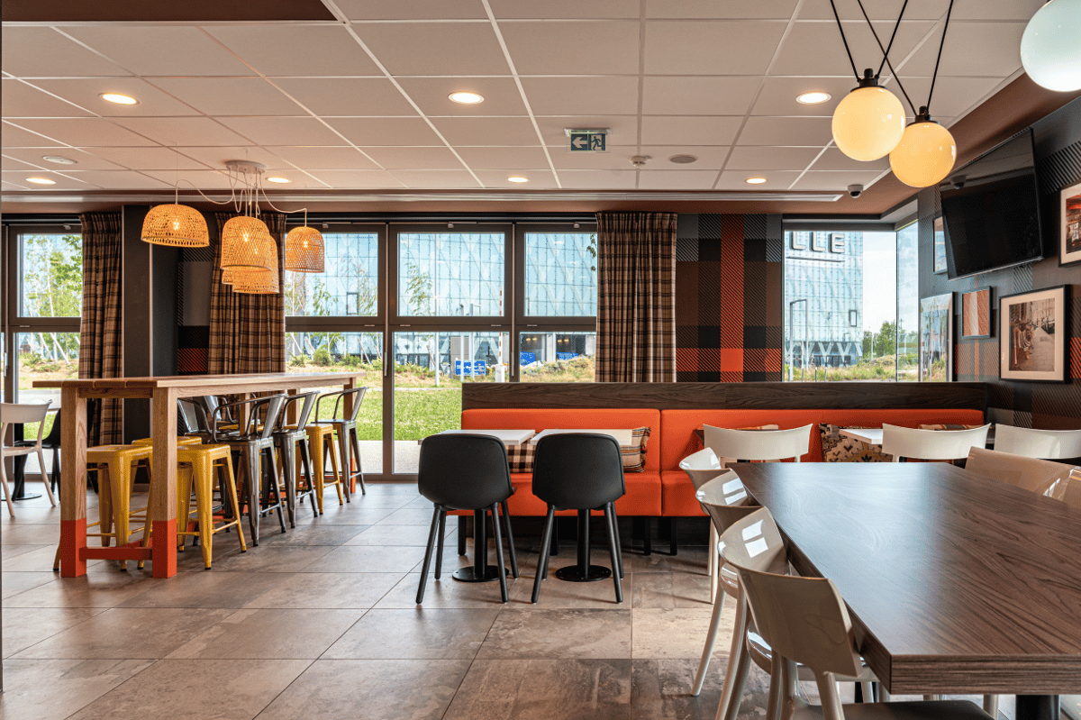 A bright, modern easyHotel CDG restaurant and dining lounge featuring communal tables with yellow stools, orange booth seating, and ample natural light, ideal for guests near Paris Charles de Gaulle Airport.