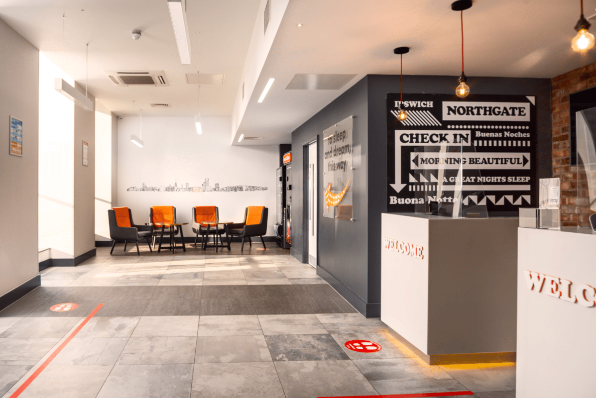 easyHotel Ipswich lobby featuring a modern reception desk, a bright seating area with orange and grey chairs, and a white wall adorned with a city skyline mural.