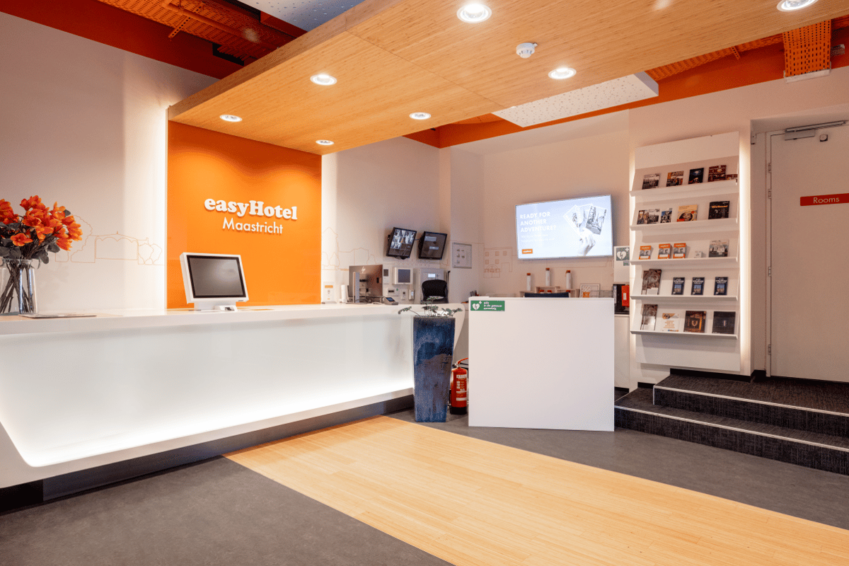 Modern reception area of easyHotel Maastricht with a sleek white desk, bright orange branded wall, and a digital display screen.