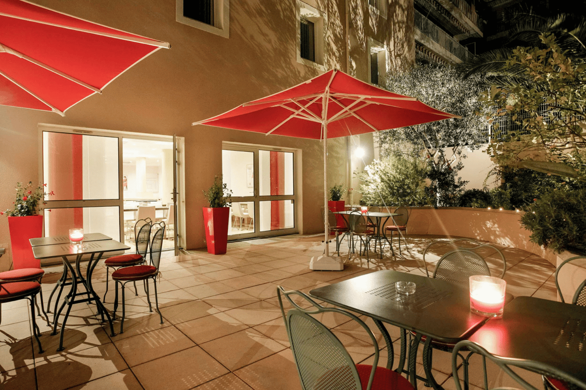 Night view of the easyHotel Nice Palais des Congres outdoor patio, featuring multiple tables, chairs with red cushions, and large red umbrellas under warm lighting, perfect for evening relaxation.