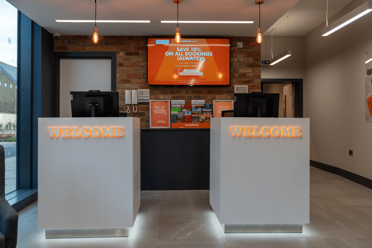 The modern reception area of easyHotel Oxford, featuring two white desks with glowing 'WELCOME' signs, a brick accent wall, and a large digital screen displaying easyHotel branding.