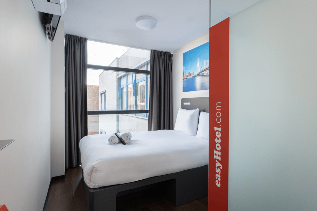 Bright easyHotel room in Rotterdam City Centre featuring a comfortable bed with white linen, two rolled towels, and a large window. A vibrant blue print of the Erasmus Bridge and Rotterdam skyline hangs above the bed.