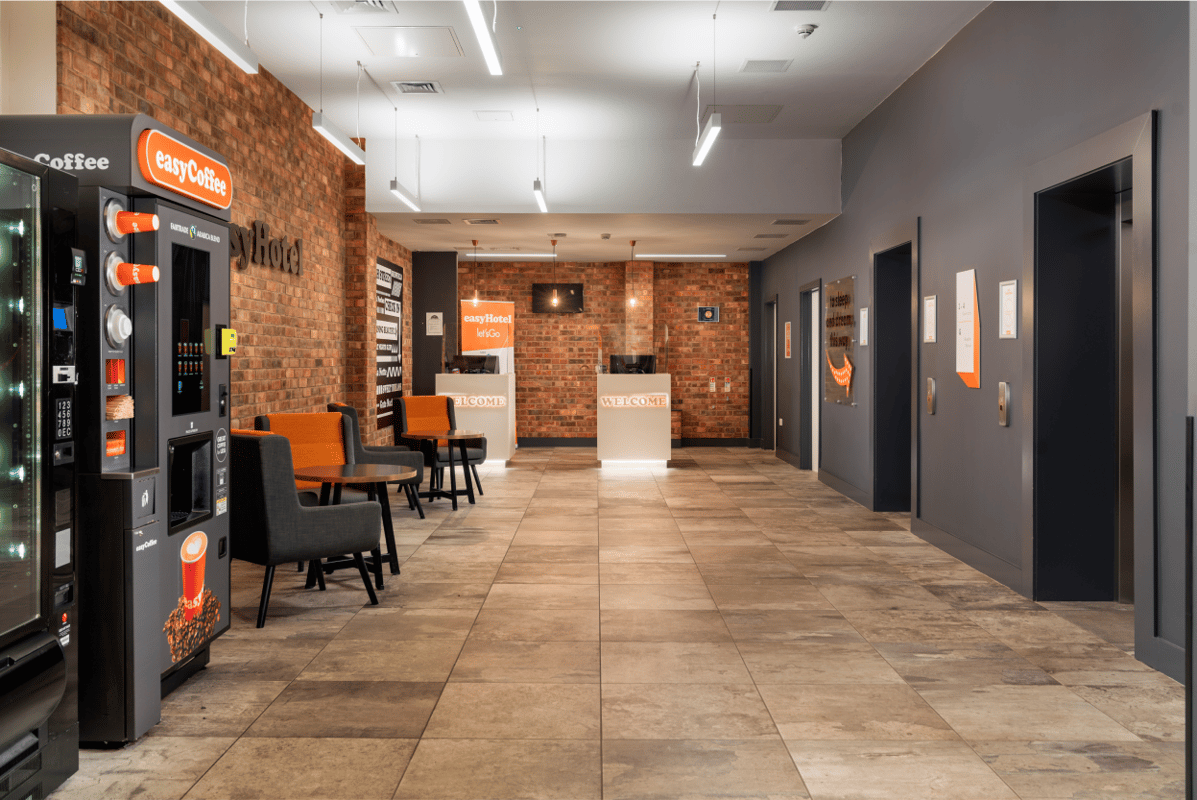 Lobby of easyHotel Sheffield featuring self-check-in kiosks, an easyCoffee vending machine, and comfortable seating, with brick and dark grey walls and tiled flooring.