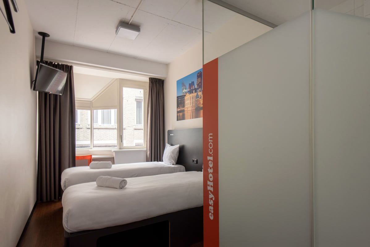 Minimalist easyHotel The Hague twin room with two single beds, city view windows, wall-mounted TV, and easyHotel branding.