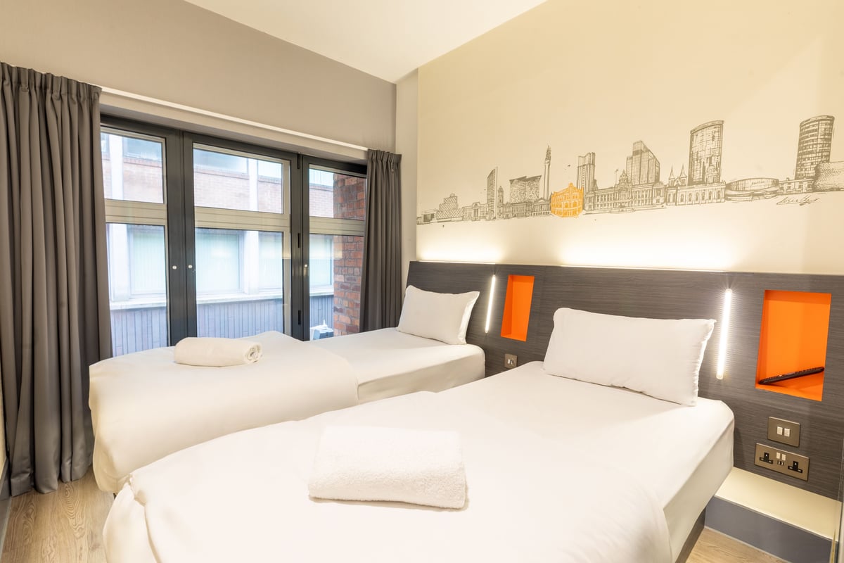 Bright easyHotel Birmingham twin room with two single beds, white linens, a dark headboard with orange accents and bedside lights, and a mural of the Birmingham skyline on the wall. A large window with grey curtains offers city views.