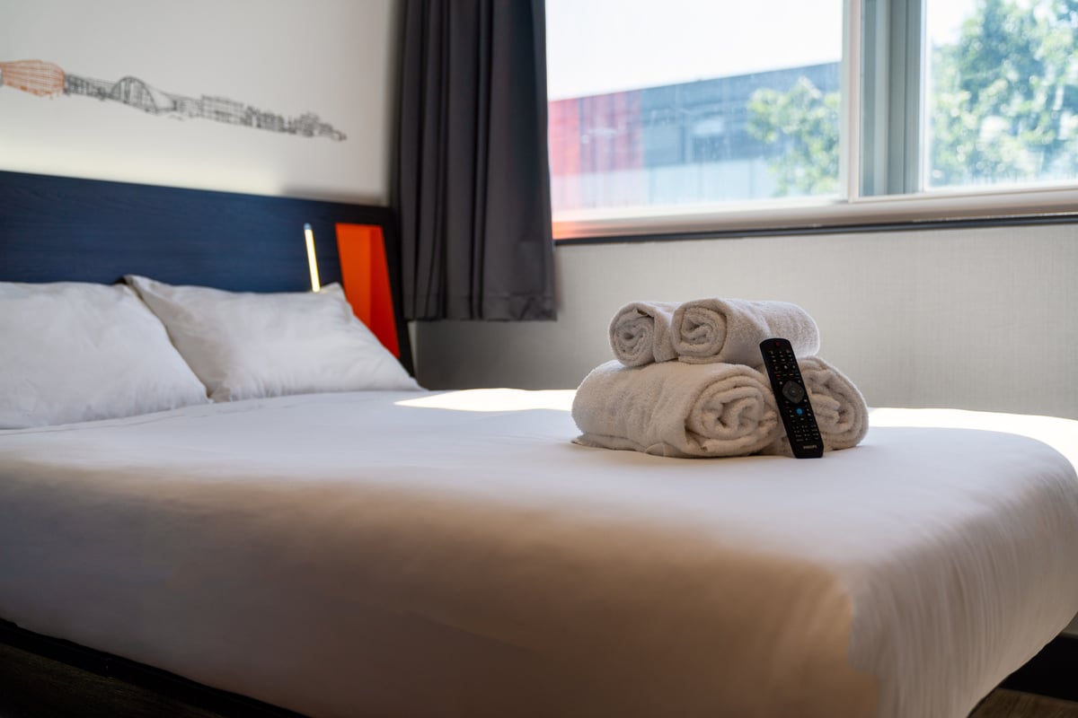Bright easyHotel Milton Keynes room with a neatly made white bed, dark blue headboard with orange accents, and rolled white towels with a TV remote on the duvet. Natural light fills the minimalist space.