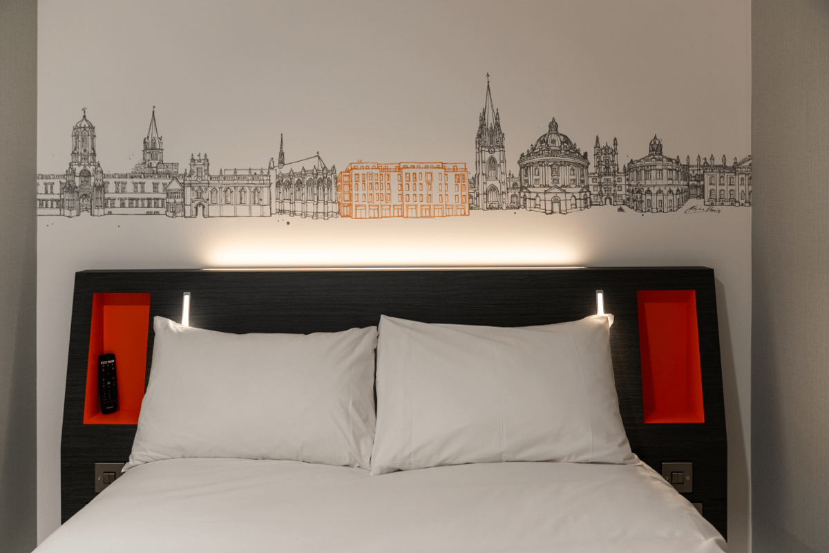 EasyHotel Oxford room featuring a comfortable double bed with white linen. Above the headboard, a detailed black and white skyline mural of Oxford's iconic buildings, including Christ Church and Radcliffe Camera, with a modern building highlighted in oran