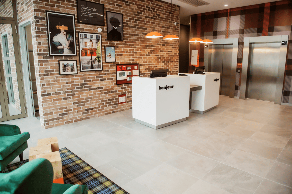 Modern easyHotel Paris Nord Aubervilliers lobby featuring a white reception desk with 'bonjour', a brick accent wall with artwork, orange pendant lights, and two elevators. Green velvet armchairs and a plaid rug are in the foreground.