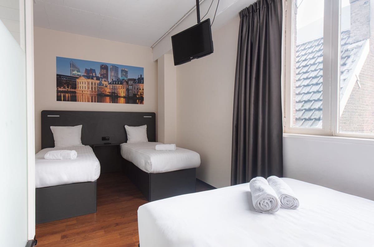 A bright easyHotel room in The Hague featuring three single beds with crisp white linen, dark grey bed frames, and a large wall mural depicting The Hague's city skyline at night.